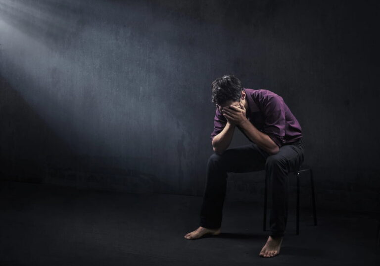 Image is of a distressed man sitting alone with his face covered in a dark room, concept of sex crime allegations and the emotional impact false accusations may have on an individual.