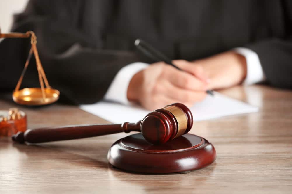 Image is of a courtroom gavel with legal scales and a lawyer writing in the background, concept of a felony DUI lawyer handling serious DUI charges in Clinton.