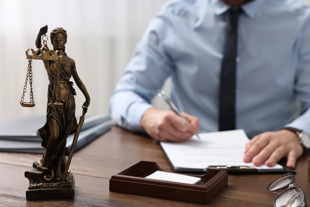 Image is of an attorney reviewing legal paperwork beside a Lady Justice statue, concept of murder defense lawyer