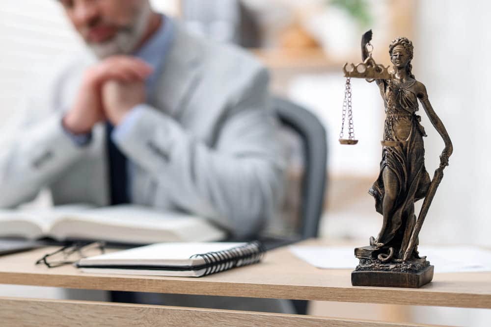 Image is of a justice statue placed in front of a lawyer reviewing legal documents in an office, concept of assault charges and building a criminal defense strategy after an arrest.