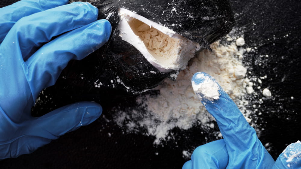Image is of blue gloved hands examining a bag containing white powder, concept of suspected controlled substance evidence in a drug manufacturing charge in Clinton.