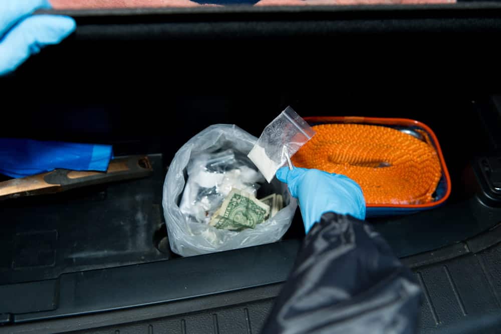 Image is of an investigator collecting a suspected drug packet and cash from a vehicle, concept of evidence supporting drug distribution charges.