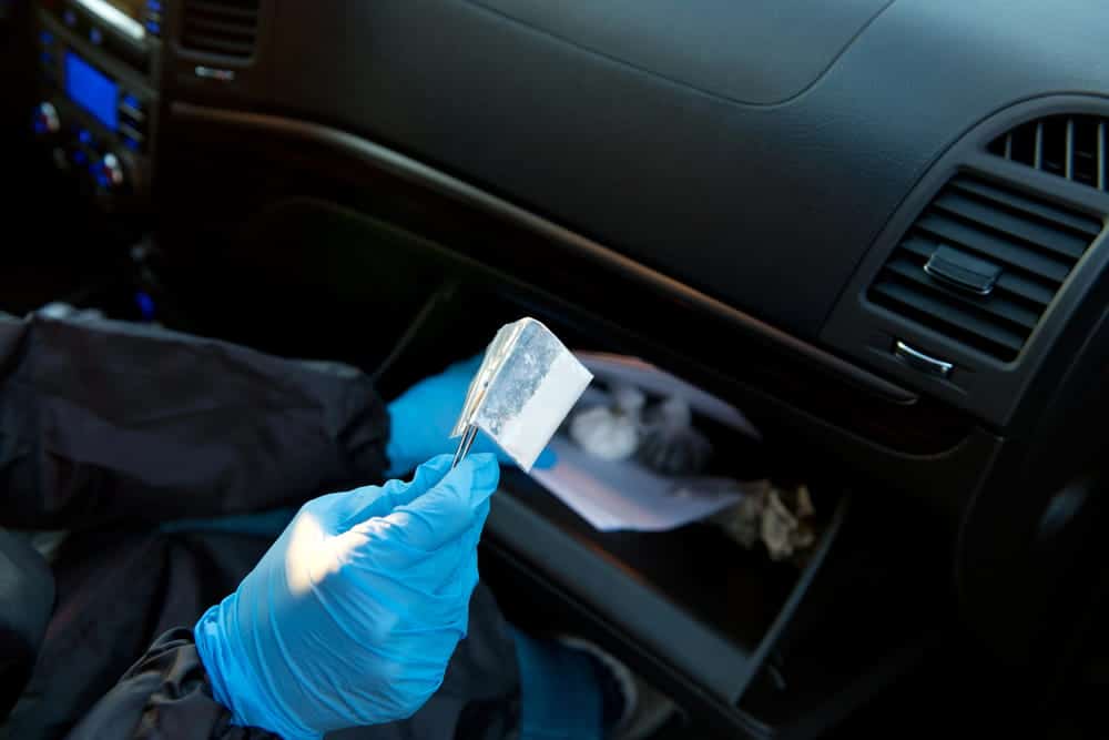 Image is of an investigator examining a suspected drug packet inside a vehicle, concept of physical evidence in drug distribution charges.